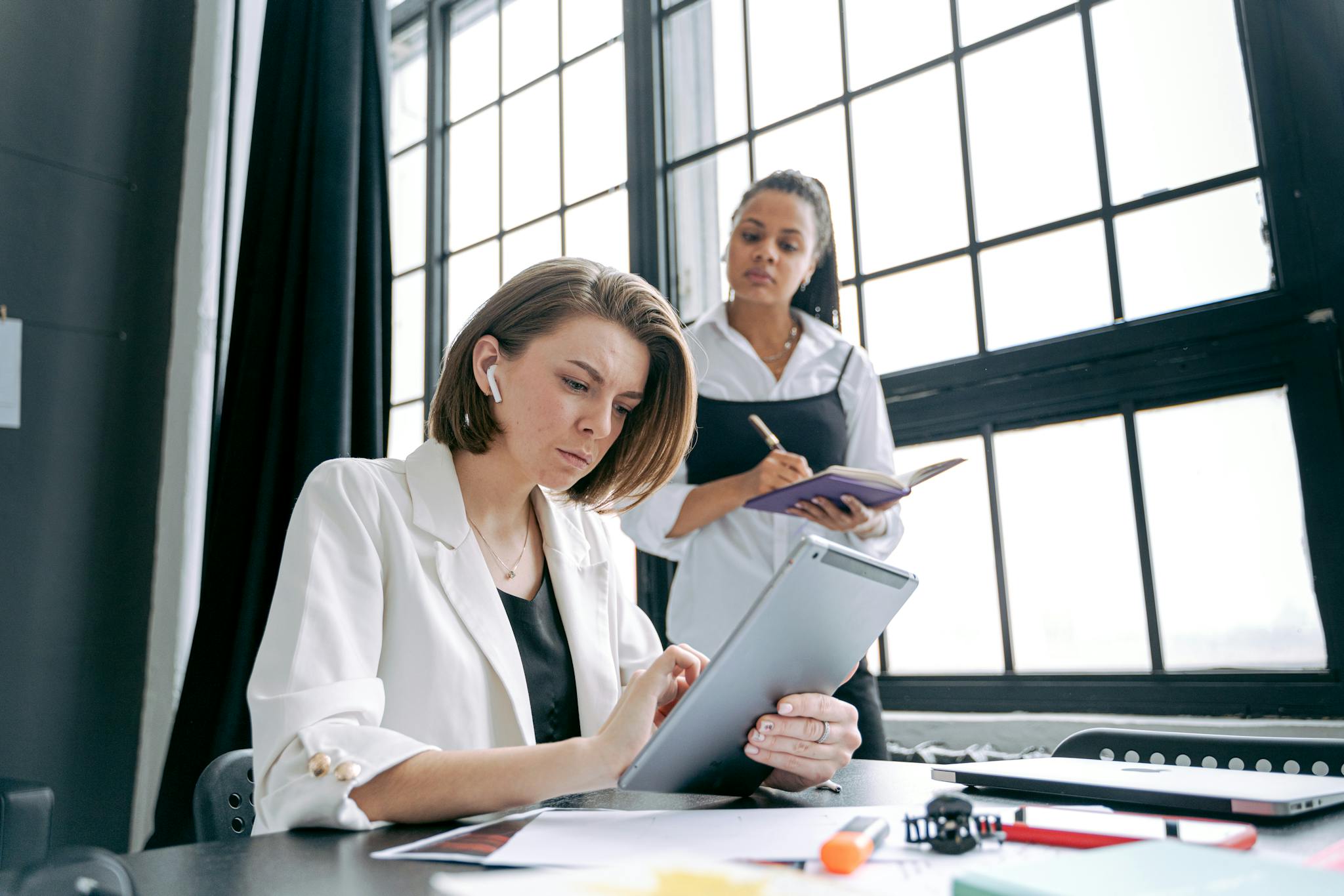 Two professional women collaborating on a project using a tablet in a modern office setting.